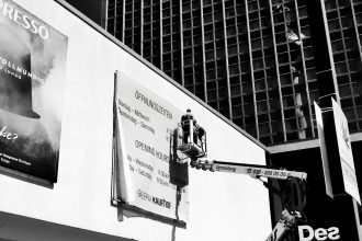 Trabalhador em uma plataforma elevatória ajusta um outdoor na fachada de um prédio.