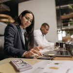 Dois profissionais analisam informações contábeis em um laptop, com uma calculadora sobre a mesa, em um escritório moderno.
