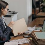 Profissional analisa documentos e arquivos em sua mesa de trabalho, ao lado de um laptop.