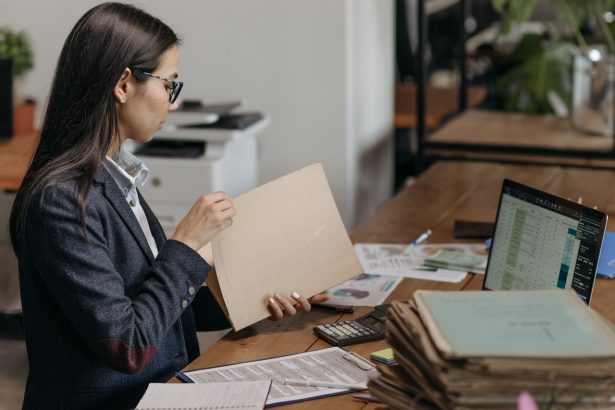 Profissional analisa documentos e arquivos em sua mesa de trabalho, ao lado de um laptop.