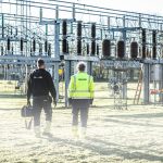 Dois trabalhadores usando equipamentos de segurança inspecionam uma subestação elétrica ao ar livre.