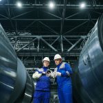 Dois trabalhadores usando equipamentos de proteção observam bobinas de aço em uma instalação industrial.
