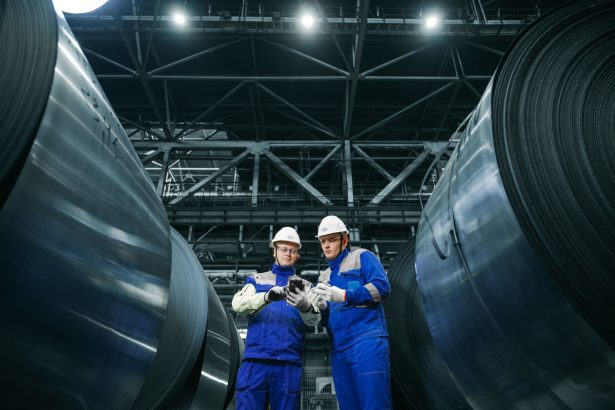 Dois trabalhadores usando equipamentos de proteção observam bobinas de aço em uma instalação industrial.