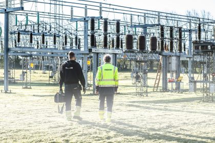Dois trabalhadores usando equipamentos de segurança inspecionam uma subestação elétrica ao ar livre.