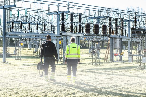 Dois trabalhadores usando equipamentos de segurança inspecionam uma subestação elétrica ao ar livre.